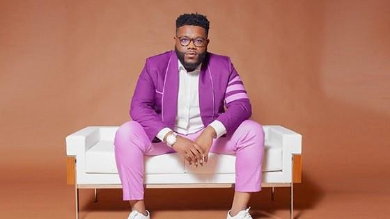 A man with black hair and a beard wearing glasses, a purple suit, and a white button down shirt while seated on a white couch against a brown background.